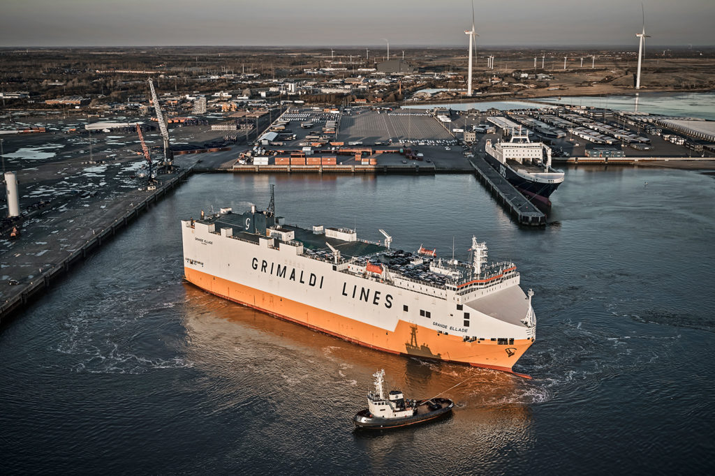 Grimaldi Lines' Grande Ellade being maneuvered by a tugboat in the harbor. In the background, a large DFDS ship and terminal areas are visible, including both the auto terminal and the multi-terminal.
